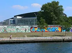 Frontale Farbfotografie von einem Flussufer mit einer Uferpromenade und einer langen Mauer mit Graffitis. Ein Mann sitzt links am Ufer und an der Mauer laufen einige Menschen. Rechts stehen vor und hinter der Mauer Bäume. Hinter der Mauer ist ein großes, modernes Glasgebäude.