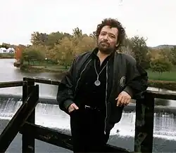 Bernard Vitet an einem Geländer gelehnt. Im Hintergrund ein Wasserfall. Er trägt eine schwarze Hose, schwarzes Hemd und eine offene schwarze Jacke. Seine rechte Hand steckt in seiner Hosentasche.