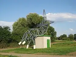 Skulptur, die einem Freileitungsmasten nachempfunden ist, der sich zum "Ausruhen" "hingesetzt" hat
