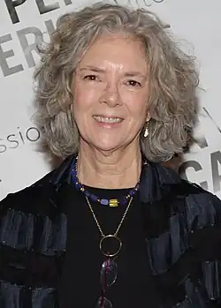 Beth Gutcheon (2014) mit grauem, lockigem Haar, die eine dunkle Kleidung trägt, lächelt freundlich. Sie trägt eine Halskette mit blauen und goldenen Perlen, und eine Brille hängt an einer Kette um den Hals. Der Hintergrund besteht aus einem Banner, auf dem teilweise lesbare Textfragmente zu sehen sind, darunter die Wörter „PER“ und „SSION“.