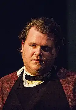 Björn Meyer trägt ein weißes Hemd mit hochgekrempelten Ärmeln, dazu eine schwarze Fliege. Über dem Hemd ist eine dunkle Schürze zu sehen, die über einem roten, gemusterten Oberteil getragen wird. Der Hintergrund ist dunkel gehalten, wodurch der Fokus auf die Person gelenkt wird. Es gibt keine zusätzlichen Objekte oder Details, die ablenken. Das Foto entstand 2015 bei einer KARLMAYSELF-Vorstellung in Hamburg.