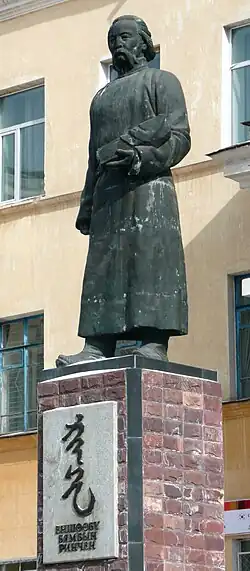 Statue in einem Grauton von Byambyn Rinchen vor einem Gebäude in lebensgroß und landestypische Bekleidung von leicht seitlich nach links schauend auf einem Sockel aus roten Steinen stehend mit einer dort weiß marmorierten Platte, die den Namen und zwei Symbole enthält.