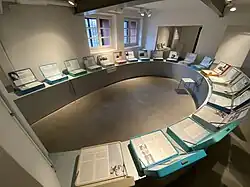 Blick in einen Raum der Dauerausstellung in der Gedenkstätte Bullenhuser Damm, auf der in Draufsicht farbige Koffer zu sehen sind, die Biografien enthalten.