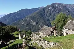 Ein grünes, tiefes Tal mit Felswänden auf der gegenüberliegenden Talseite und einem winzigen Dörfchen unterhalb mehrerer Berggipfel im bewaldeten Hang. Im Vordergrund Steinhäuser ohne Putz mit groben Steindächern (Rustico) und ein steiler Bergwanderweg zwischen hellgrünen terrassierten Almwiesenflächen. Darüber ein blauer Himmel.