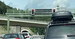 Eine Schilderbrücke quer über einer Straße. In der Mitte der Brücke ist eine leuchtende Anzeige „Blockabfertigung“ angebracht, rechts und links davon je eine rot zeigende Ampel. Unter der Brücke stauen sich einige Autos. Im Hintergrund ist ein Tunnelportal zu erkennen.