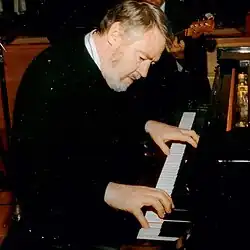Bob Thompson spielt Klavier, konzentriert und mit einem leichten Lächeln, in einem musikalischen Umfeld. Er trägt einen schwarzen Pullover über einem weißen Hemd. Seine Hände sind auf den Tasten des Klaviers positioniert, die Finger in einer Spielhaltung. Im Hintergrund erkennt man eine weitere Person, die ein Saiteninstrument spielt, vermutlich eine Gitarre oder einen Bass. Die Szene scheint in einem Innenraum aufgenommen worden zu sein, möglicherweise während einer musikalischen Darbietung oder Probe.