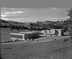 Ein Schulhaus im brutalistischen Stil steht an einem Wiesenhang idyllischer Landschaft.