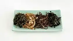 Auf einer weißen Platte sind vier Gemüsehäufchen drapiert, erstens ein grünes Blattgemüse, zweitens sesambestreute helle Rettichstreifen, drittens bräunliche Adlerfarn-Schösslinge und viertens ein dunkleres grünes Blattgemüse.