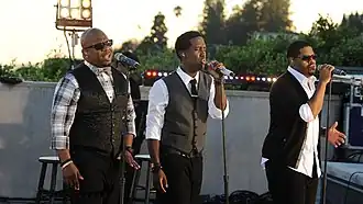 Boyz II Men stehen vor drei Mikrofonen und singen. Sie tragen schwarze Hosen sowie (von links nach rechts) ein schwarz-grau kariertes Hemd mit grauer Fliege und schwarzer Weste; ein weißes Hemd mit schwarzer Krawatte und grauer Weste; ein weißes Hemd mit schwarzem Jackett. Im Hintergrund sind eine graue Wand, Bühnenbeleuchtungsanlagen und eine Hecke zu sehen.