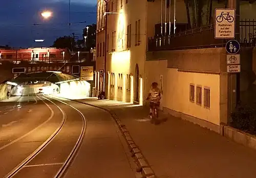 Eine lange Bahnunterführung in Augsburg mit leichtem Gefälle, glänzende Meterspur-Straßenbahnschienen mit etwa 1,50 m Abstand vom Bordstein, Überholverbot (durchgezogene weiße Linie) für Autos in der beleuchteten Unterführung nahe dem Hauptbahnhof mit einem rot-weißen Zug oberhalb der leicht gebogenen Tunnelüberwölbung. Eine Fahrradfahrerin (vermutlich) auf dem etwa 2,50 Meter breiten Gehweg rechts, ein Bettler sitzt ein wenig weiter unten am Rand an der hohen Hauswand. Verkehrsschild Fussgänger mit Zusatzschild Fahrradfahren frei und Hinweistafel „Radfahren auf der Fahrbahn erlaubt“. Links auf der gegenüberliegenden Straßenseite trennt keine Wand mehr den Bürgersteig von der aufwärtsführenden Fahrbahn ab. Blaue Stunde am Abend mit Gebäude einer Brauerei rechts im Bild.