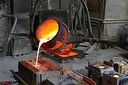 Aus einem rot glühenden Eimer, der an einer Stange befestigt und leicht nach vorne geneigt ist, fließt gelb glühende flüssige Bronze in ein Loch einer rechteckigen Form.