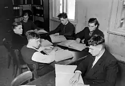 Fünf junge Männer und eine Frau sitzen um einen Tisch mit den Händen auf großen, hellen aufgeschlagenen Büchern, vermutlich mit Blindenschrift. Ein Stuhl mit gebogener Holz-Rückenlehne ist noch frei. Im Hintergrund ein Bücherregal in der Zimmerecke und ein helles, zweiflügliges Fenster.