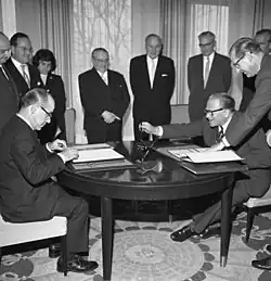 Schwarzweißfoto von neun Männern und einer Frau, die formell gekleidet sind. Zwei sitzen sich an einem runden Tisch gegenüber: links der spanische Botschafter Luis de Urquijo, rechts der deutsche Staatssekretär Hilger van Scherpenberg, Dokumente in aufgeschlagenen Mappen vor sich. Ein Mann beugt sich über den rechts Sitzenden, alle anderen stehen im Hintergrund und sehen zu. Scherpenberg streckt seinen Arm über den Tisch, einen Gegenstand in der Hand. Durch die Fenstergardinen sind Umrisse von Bäumen erkennbar.