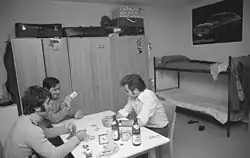 Auf dem Schwarzweißfoto ist ein Raum zu sehen, mit einem Etagenbett, drei Kleiderschränken, einem halbhohen Kühlschrank und einem Tisch, an dem drei Männer sitzen, die Karten spielen. Auf dem Tisch sind zwei Bierflaschen und -gläser, ein Wecker und diverse andere Gegenstände. Auf den Kleiderschränken liegen mehrere Koffer, über dem Etagenbett hängt ein Poster, auf dem ein Auto abgebildet ist. Die Männer tragen informelle Kleidung und Armbanduhren. Sie sind in ihr Spiel vertieft und wirken vergnügt.