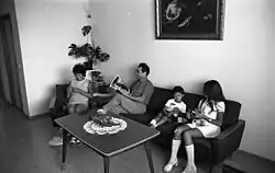 Auf diesem Schwarzweiß-Foto ist eine Sitzecke in einem Wohnzimmer mit (von links nach rechts) einer Frau auf einem Sessel sowie einem Mann, einem kleinen Jungen und einem Mädchen im Teenageralter auf einem dunken Sofa. Sie sind im Stil der 1950er-Jahre gekleidet und jeweils mit einem Gegenstand, den sie in Händen halten, beschäftigt. Zum Beispiel ist der Mann anscheinend dabei, eine Zeitschrift zu lesen. Auf dem Couchtisch vor ihnen liegt ein Stickdeckchen mit einem Gegenstand darauf. An der Wand hinter dem Sofa hängt ein gerahmtes Gemälde, ansonsten sind die Wände weiß. In der Ecke steht ein Blumenständer mit großblättrigen Zimmerpflanzen.