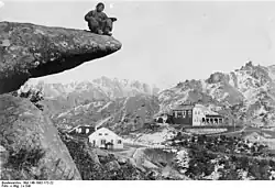 Im Vordergrund des schwarz-weiß Bildes ist links ein Felsvorsprung mit einer sitzenden Person zu sehen. Im Hintergrund sind verschiedene Gebäude in einer hügeligen Landschaft erkennbar.