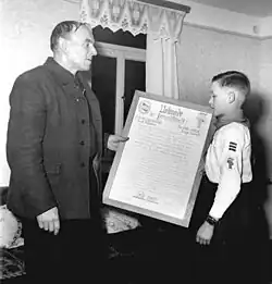 Ein Pionier mit drei Streifen am Ärmel des Pionierhemdes, die ihn als Vorsitzenden des Freundschaftsrats kennzeichnen, überreicht einen Freundschaftsvertrag an die Patenbrigade