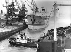 Schwarz-Weiß-Foto eines Hafens, mehrere große Schiffe, eines wird links von einem Hafenkran be- oder entladen. Von der MS Zwickau ist nur ein Teil zu erkennen, wohl weil das Foto von dem Schiff aus gemacht wurde. Vorne ist außerdem ein Schlepperboot zu sehen, dass möglicherweise die Zwickau abschleppt. Hinten ist durch Nebel nicht viel zu erkennen.