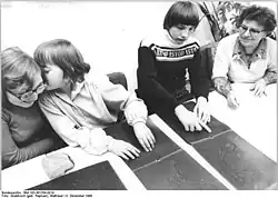 Das Schwarzweißfoto aus dem Jahre 1980 zeigt drei Jugendliche und eine erwachsene Frau an einem Tisch sitzend. Auf diesem liegen Bücher für Blinde, auf deren dunklen Seiten reliefartig, also tastbar, Tierdarstellungen (Löwe, Giraffe) eingeprägt sind. Links haben zwei Mädchen die Köpfe zusammengesteckt und tuscheln vergnügt miteinander. Die rechte der beiden tastet dabei mit geschlossenen Augen mit den Fingerspitzen beider Hände über eine Buchseite, während sie der neben ihr Sitzenden ins Ohr flüstert. Diese, mit einer dicken Brille ausgestattet, hört belustigt zu. Der daneben sitzende, langhaarige Junge ist offenbar blind; er betastet konzentriert das vor ihm liegende Buch. Die Betreuerin schaut zu.