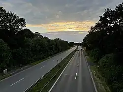Blick auf eine autobahnähnlich ausgebaute Bundesstraße von einer Überführung aus. Links und rechts der Fahrbahn stehen Büsche und Bäume, in der Mitte zwischen den je zwei Fahrstreifen pro Richtung befindet sich ein mit Leitplanken gesicherter Mittelstreifen. Es sind im Hintergrund ein Auto und ein Wohnwagen auf den beiden rechten Fahrstreifen erkennbar. Im Vordergrund stehen Radarfallen („Blitzer“). Über der Straße ein dramatischer Wolkenhimmel (Abenddämmerung).