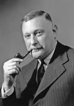 Mann im mittleren Alter im Seitenprofil nach links vorne schauend im grauen Jackett, hellem Hemd und dunkler Krawatte sowie eine Pfeife rauchend in schwarzweiß.