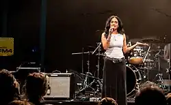 Eine junge Frau mit dunklen Haaren, und braunen Augen. Sie hält ein Mikrofon in der Hand, hat ein weißes Top, einige Ketten, Ringe, Ohrringe und einen glänzenden großen Gürtel dazu einen Schwarzen Rock, sie steht auf einer Bühne, hinter ihr ist ein Schlagzeug zu sehen und ein Radio FM4 Logo, vor ihr eine Menge von Menschen, die sie anschauen.