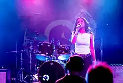 Chovo eine junge Frau mit dunklen Haaren und Augen steht auf einer Bühne. Sie trägt ein weißes Top, schwarzes Kleid, viele silberne Ringe und Ohrringe, sowie einen glänzenden Gürtel. Sie hält ein Mikrofon zum Mund und singt. Bühnenlichter beleuchten Sie in den Farben Rosa und den Hintergrund in Blau. Im Vordergrund sieht man ein paar Menschen aus dem Publikum. Im Hintergrund steht ein Schlagzeug und ein paar Gitarrenverstärker