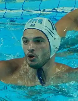 Kolomvos ist im Wasser, er trägt eine weiße Kopfbedeckung.