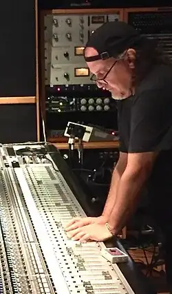 Claudio Cueni arbeitet an einem Mischpult in einem Tonstudio. Er trägt ein schwarzes T-Shirt sowie eine schwarze Mütze, unter der lange Haare hervorschauen. Er beugt sich konzentriert über das Mischpult und hat beide Hände darauf gelegt. Das Mischpult selbst ist mit vielen Reglern und Knöpfen ausgestattet, die dazu dienen, Tonaufnahmen und -mischungen zu steuern. Im Hintergrund sind verschiedene technische Geräte und Regler zu sehen, die ebenfalls für die Bearbeitung von Tonmaterial verwendet werden.