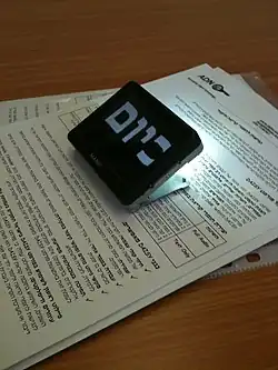 Auf einem Tisch liegen einige Blätter mit hebräischem Text, darauf wurde das etwa zigarettenschachtelgroße Lesegerät platziert. Mit einer kräftigen Lichtquelle beleuchtet es einen kleinen Teil des Textes, auf seiner Oberseite erscheinen wenige hebräische Schriftzeichen in sehr starker Vergrößerung (mehrere Zentimeter hoch).