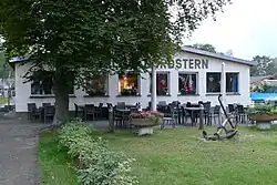 Breites Haus hinter Bäumen, davor Terrasse mit Stühlen und Tischen und ein Anker auf Grasfläche