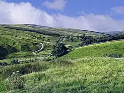 Blick von einem seichten mit Wiese bewachsenen Erhebung auf eine zweite sehr langgezogene schräge Ebene, welche weit ins Land geht und von einer kleinen Straße langsam und mit langen Kurven überzogen wird