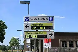 Blick auf drei längliche weiße Schilder. Ganz oben mit schwarzer Schrift „Parkleitsystem“, auf beiden Seiten ein blaues Rechteck mit einem weißen P unter einem weißen Dach. Das Schild darunter weist mit einem schwarzen Pfeil nach links, daneben steht auf gelbem Grund „Rheinufer“. Unter diesem schwarzen Schriftzug ist eine rechteckige schwarze Anzeige angeordnet. Sie zeigt mit einer blockigen hellgelben Schrift „211 FREI“ an. Das Schild darunter ist gleich aufgebaut, weist aber nach rechts. Die Schrift lautet oben „Rheinterrasse“, unten „22 FREI“. Hinter den Schildern erkennt man den Himmel, einige Bäume und Gebäude.