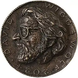 Vorderseite einer Bronzemedaille mit dem kunstvoll gestalteten Porträt von David Wigg-Wolf in hohem Relief. Das plastisch hervorgehobene Bildnis wird von einer kreisförmigen Inschrift mit dem Namen ‚DAVID WIGG-WOLF‘ und dem Jahr ‚2024‘ umgeben.