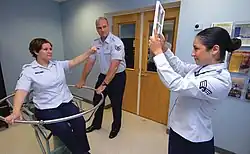 Eine Versuchsperson sitzt in einem Drehstuhl. Eine zweite Person hat gerade die Rotation des Stuhls gestoppt. Die orientierungslose Versuchsperson zeigt auf ein Zeichen, welches von einer dritten Person gehalten wird.