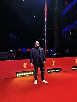 Denis „Marshall“ Ölmez, der auf dem roten Teppich der Berlinade 2023, steht. Er trägt ein dunkles Sakko, ein schwarzes T-Shirt, dunkle Jeans und weiße Turnschuhe. Der Hintergrund des Bildes ist dunkel gehalten, mit einigen hellen Lichtern und Reflexionen. Eine rote Absperrung mit dem goldenen Logo der Berlinale, einem stilisierten Bären, ist sichtbar.