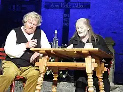 Links sitzt der Brandner Kaspar in beiger Hose, weißem Hemd und schwarzer Weste mit einem Schnapsglas in der rechten Hand. Der linke Ellenbogen lehnt auf dem Holztisch im Zentrum des Bildes. Der Tisch ist von schräg unten fotografiert, man kann aber die Schnapsflasche erkennen, ein Tischlicht und ein Glas vor der zweiten Person, dem Boanlkramer. Dieser ist schwarz gekleidet, das Gesicht ist weißgeschminkt. Er hat einen Haarkranz mit langen Haaren rund um die kahle Kopfmitte. Im Hintergrund ist ein Kreuz zu sehen mit der Aufschrift „Zum Heile Aller“ und den Jahreszahlen 1961, 1972 und 1990.