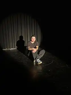 schwarze Bühne in der Kulisse Wien. Der Kuseng sitzt auf dem Boden und ein Scheinwerferlicht leuchtet ihn an.