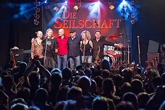 Sechs Musikanten in einer Reihe am vorderen Bühnenrand, vorne Publikum, hinten ein Banner mit der Aufschrift „Die Seilschaft“.