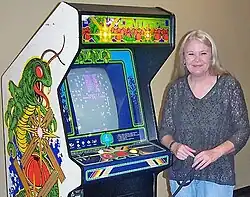 Dona Bailey steht neben einem Arcade-Automaten, auf dem das von ihr entwickelte Spiel Centipede läuft.