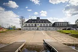 Blick über Slipanlagen und Stege hinweg auf ein weißes Gebäude. Im unteren Stockwerk sieht man (Boots-?)garagentüren.