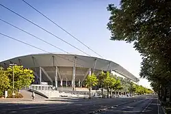 Foto eines weißen Stadiongebäudes über eine Straße mit Trambahnschienen und -leitungen hinweg.