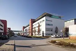 Foto eines modernen weißen Gebäudes mit rötlichen, etwas vorstehenden Zwischenstücken, auf dem ein weiß-grünes Logo und Schriftzug „Fraunhofer“ zu sehen ist. Vor dem Gebäude sind Straßen und Parkplätze zu sehen.