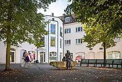 Fotos eines von Häusern gesäumten Platzes mit einer halbrunden Bankreihe um ein Denkmal einer Bronzefigur, die auf einem Stein sitzt