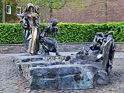Das Edith-Stein-Denkmal, einer Bronze mit den drei Figuren links und einer nach rechts führenden Rampe, die vor aufgehäuften Schuhen endet. Im Hintergrund eine leuchtend grüne Hecke