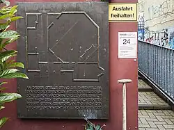 Gedenktafel mit Grundriss der Synagoge und Inschrift „An dieser Stelle stand die Ehrenfelder Synagoge, verbunden mit einer Religionsschule für Mädchen und Jungen, erbaut 1927 nach dem Entwurf des Architekten Robert Stern, zerstört am Tag nach der Reichspogromnacht am 9. November 1938“. Rote Hausfassade, neben der Gedenktafel direkt ein gelbes „Ausfahrt verboten“-Schild und ein Handzettel des körnerstrassenfests. Daneben ragt noch der Stiel eines Spatens ins Bild