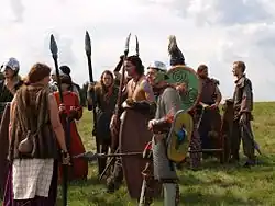 Ein Dutzend Personen in keltisch anmutender Ausstattung mit Speeren, Schilden und Schwertern stehen auf einer sonnenbeschienenen Wiese.