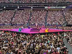 Swift steht mit ihrer Gitarre auf einer Hydraulik-Plattform auf einer pinken Bühne. Um ihr herum stehen Tänzerinnen und ihre Band.