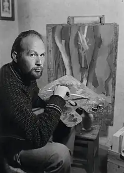 Erich Ess in seinem Atelier, 1959, mit Blick in die Kamera und einer Farbpalette in der Hand. Im Hintergrund ist ein Bild auf einer Staffelei zu sehen.