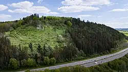 Drohnenaufnahme, vorne unten eine Bundesstraße mit wenigen Kraftfahrzeugen, dahinter erhebt sich ein mit Mischwald bewachsener kleiner Berg. Zum Gipfel mit seinen Felsen hin ist eine Fläche am Steilhang gerodet und von einem Pfad entlang des Hangs durchzogen